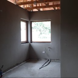 Vitalii Korneliuk - Wnętrze po tynkowaniu maszynowym: gładkie, szare ściany, drewniane okna, widoczne instalacje pod sufitem i na podłodze. Surowy stan wykończenia, przygotowany do dalszych prac.