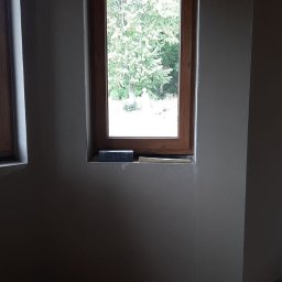Vitalii Korneliuk - Ściana po tynkowaniu maszynowym z drewnianym oknem, widoczne narzędzia i rury przy podłodze. Szare ściany, surowy stan wykończenia wnętrza.