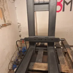 Czarne, metalowe widły do wózka widłowego, świeżo po malowaniu, stojące na drewnianych paletach w surowym pomieszczeniu z pomalowaną ścianą.