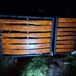 Dwuskrzydłowa brama wjazdowa z czarną ramą i poziomymi deskami z surowego drewna o nieregularnych krawędziach, widoczna w nocy obok muru z cegły i trawy, z podłączonym przedłużaczem.