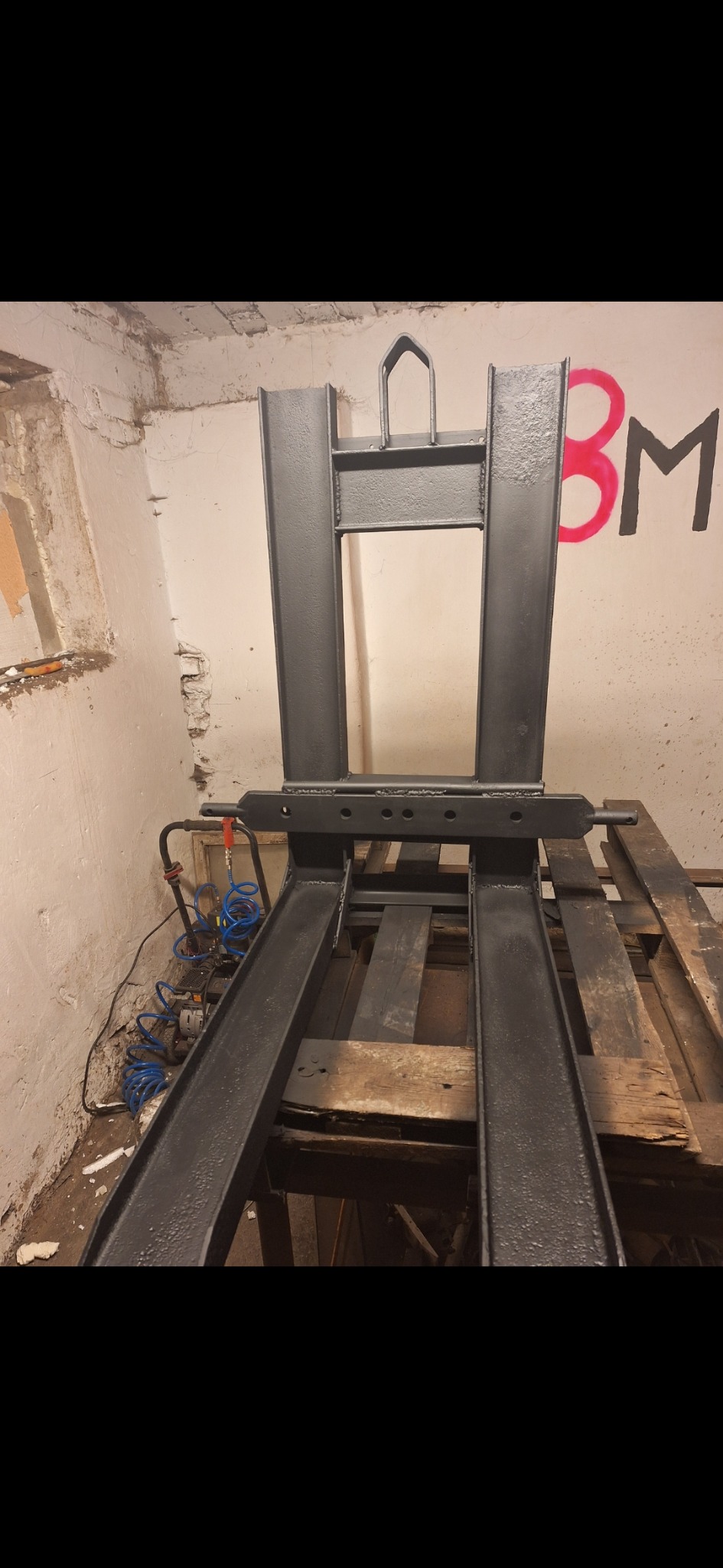 Czarne, metalowe widły do wózka widłowego, świeżo po malowaniu, stojące na drewnianych paletach w surowym pomieszczeniu z pomalowaną ścianą.