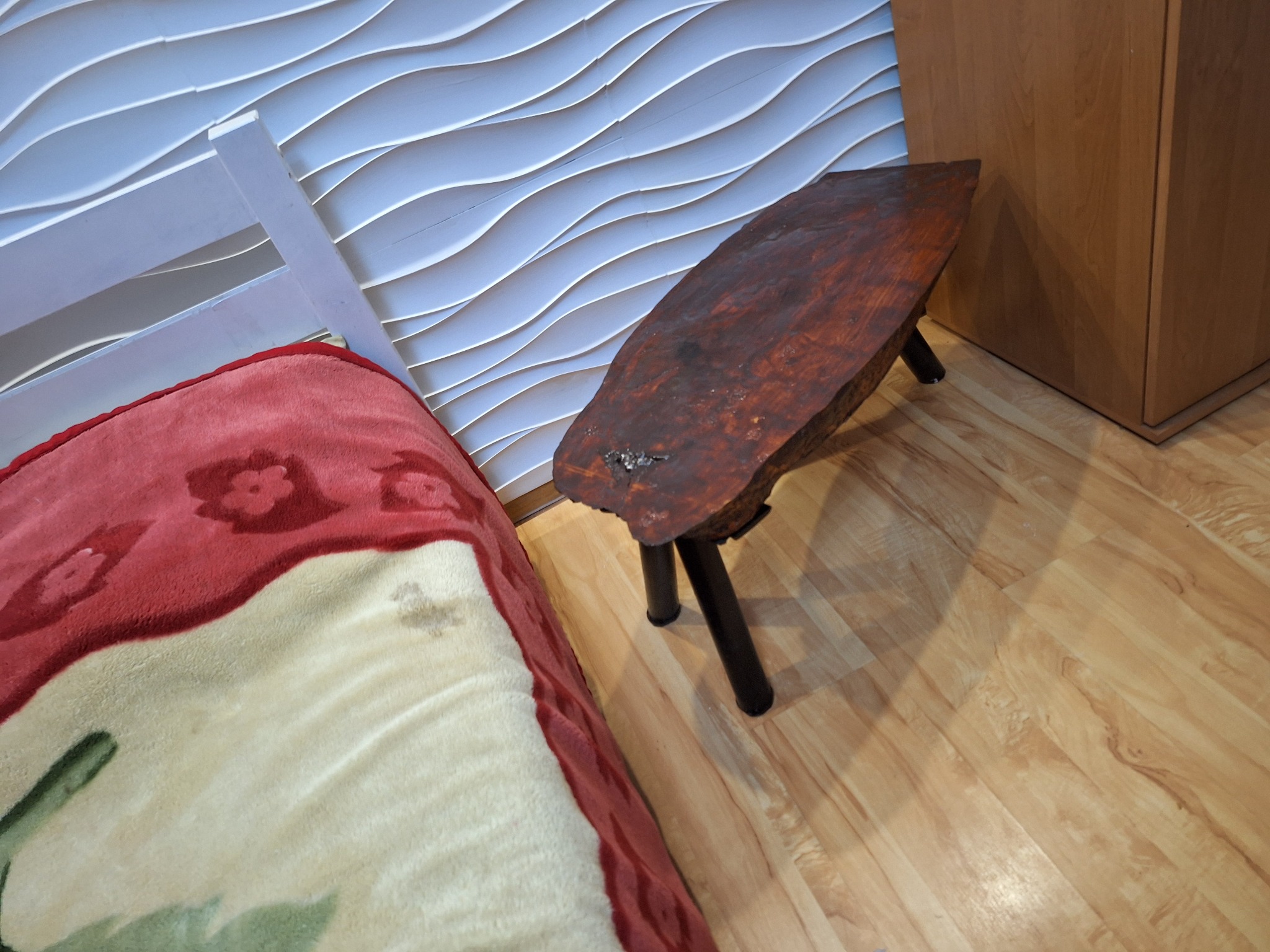 Niski, trójnożny stołek z blatem z ciemnego drewna o nieregularnym kształcie, stojący na parkiecie obok łóżka z wzorzystą narzutą i dekoracyjnej, falistej ściany.