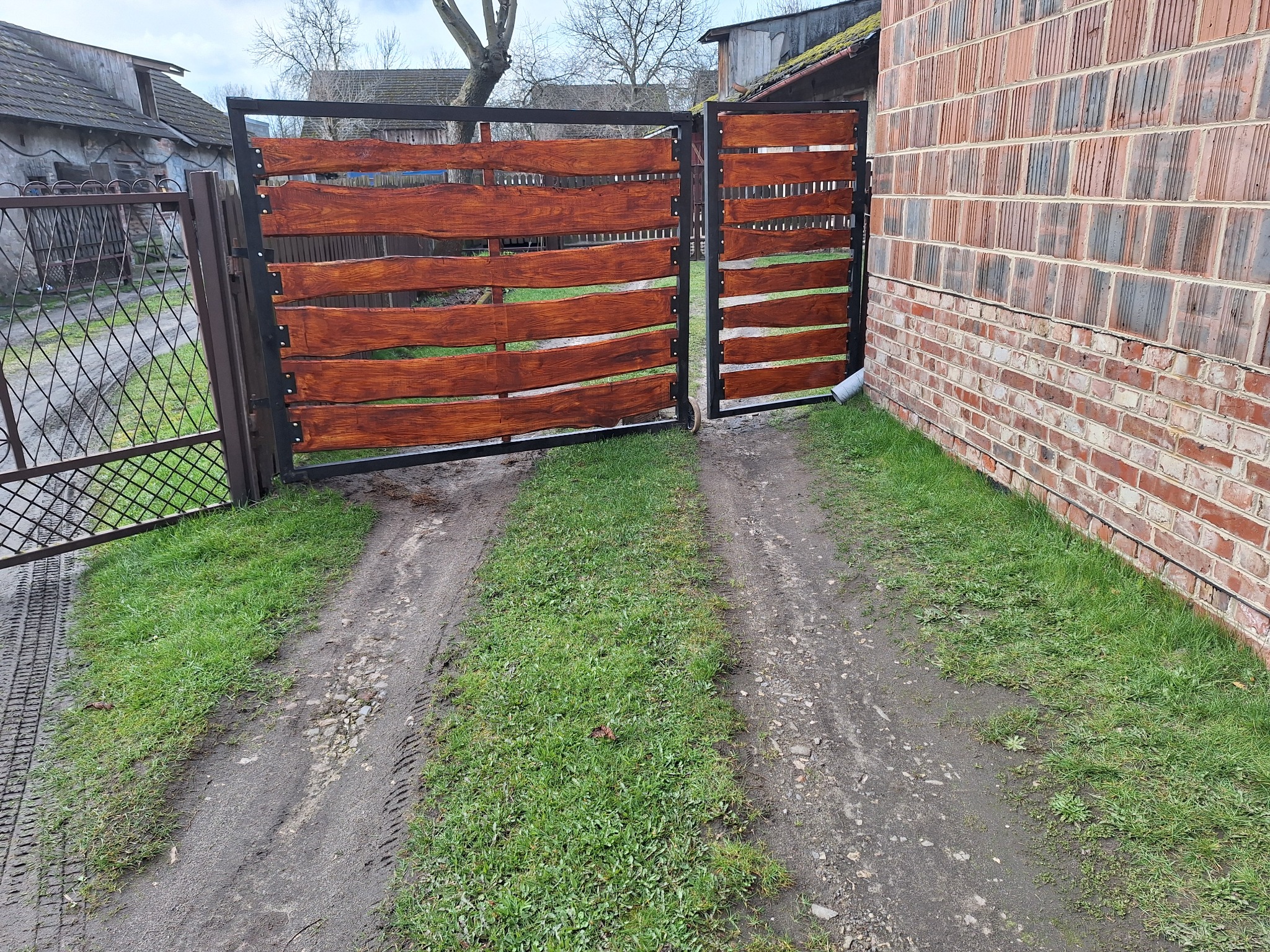 Otwarta dwuskrzydłowa brama wjazdowa z poziomymi deskami z surowego drewna w ciemnym odcieniu, osadzona w czarnej metalowej ramie, prowadząca przez trawnik i polną drogę, z budynkiem z czerwonej...