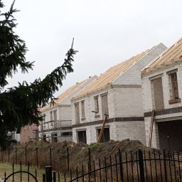 ADNN Adam Płecha - Szereg budynków w stanie surowym otwartym, wzniesionych z bloczków, z drewnianą konstrukcją dachu, widoczne zza metalowego ogrodzenia i drzewa iglastego w pochmurny dzień.