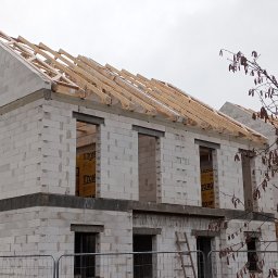 ADNN Adam Płecha - Dwukondygnacyjny budynek w stanie surowym otwartym, wzniesiony z białych bloczków, z widoczną konstrukcją dachu z drewnianych krokwi, zabezpieczony siatką ogrodzeniową.