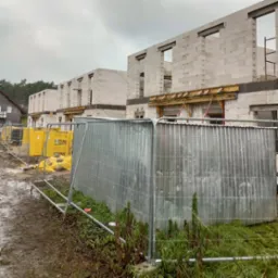 Budowa szeregowych domów z białych bloczków, widoczne pakiety materiałów budowlanych Ytong i tymczasowe ogrodzenie placu budowy w pochmurny dzień.