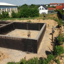 ADNN Adam Płecha - Zabezpieczone hydroizolacją ściany fundamentowe z betonu komórkowego na etapie budowy domu, z wystającymi prętami zbrojeniowymi, w tle zabudowa jednorodzinna.