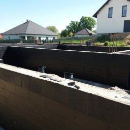 ADNN Adam Płecha - Surowe mury fundamentowe z betonu komórkowego, zabezpieczone czarną izolacją przeciwwilgociową, widoczne wiadra z preparatem i narzędzie murarskie, w tle dwa domy jednorodzinne.