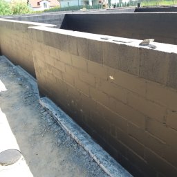 ADNN Adam Płecha - Świeżo zaizolowane, pomalowane na czarno mury fundamentowe z bloczków betonowych, widoczne narzędzia murarskie na górnej krawędzi, w tle zabudowa jednorodzinna.