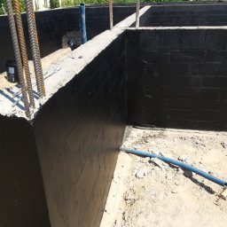 ADNN Adam Płecha - Świeżo zaizolowane, czarne ściany fundamentowe z widocznym zbrojeniem i niebieską rurą karbowaną na ziemi.