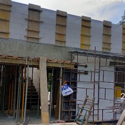 ADNN Adam Płecha - Wznoszenie ścian domu z betonu komórkowego, widoczne szalunki, rusztowania i podpory tymczasowe, teren budowy w otoczeniu drzew.