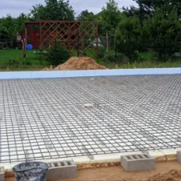 Przygotowany fundament z izolacją styropianową i siatką zbrojeniową, widoczne bloczki betonowe, w tle ogr&oacute;d z tunelem foliowym.