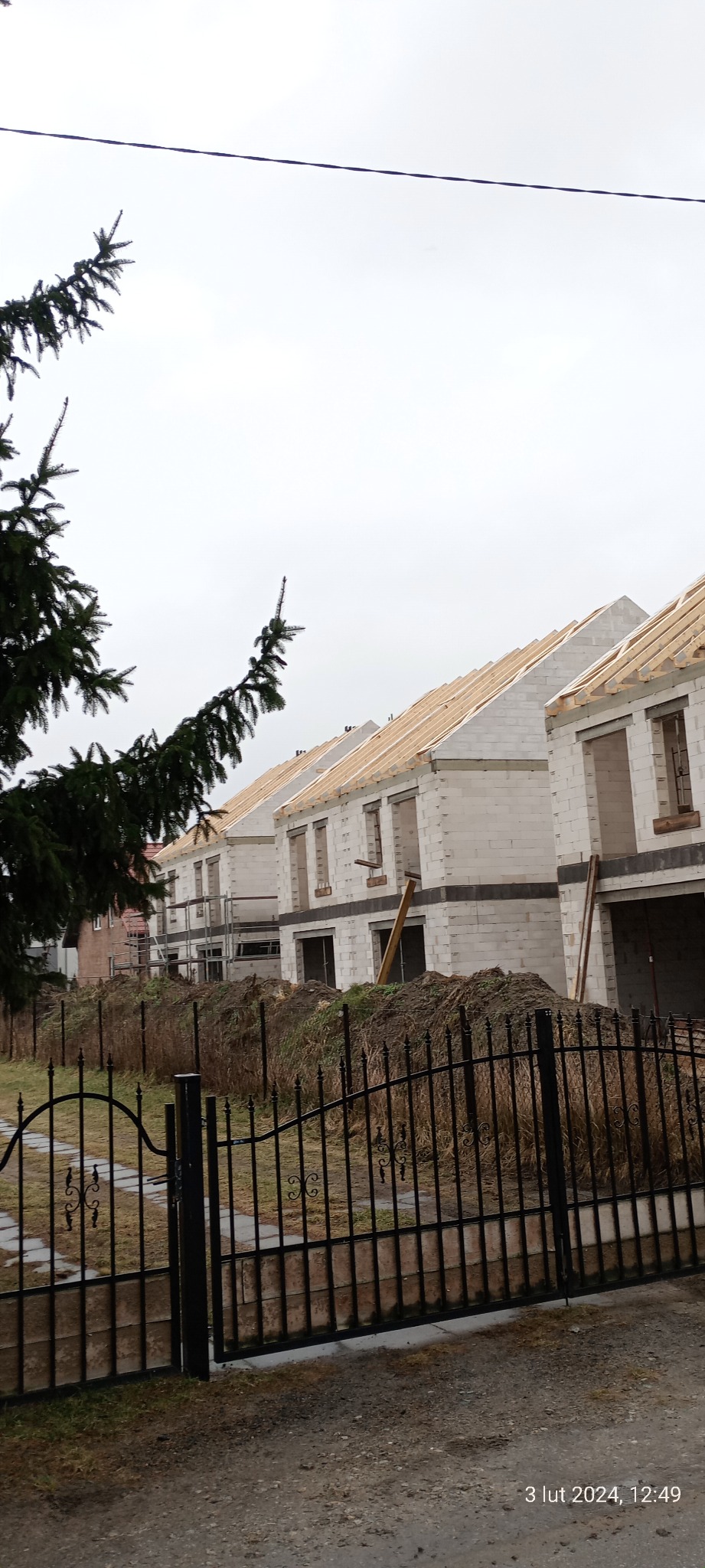 Szereg budynków w stanie surowym otwartym, wzniesionych z bloczków, z drewnianą konstrukcją dachu, widoczne zza metalowego ogrodzenia i drzewa iglastego w pochmurny dzień.