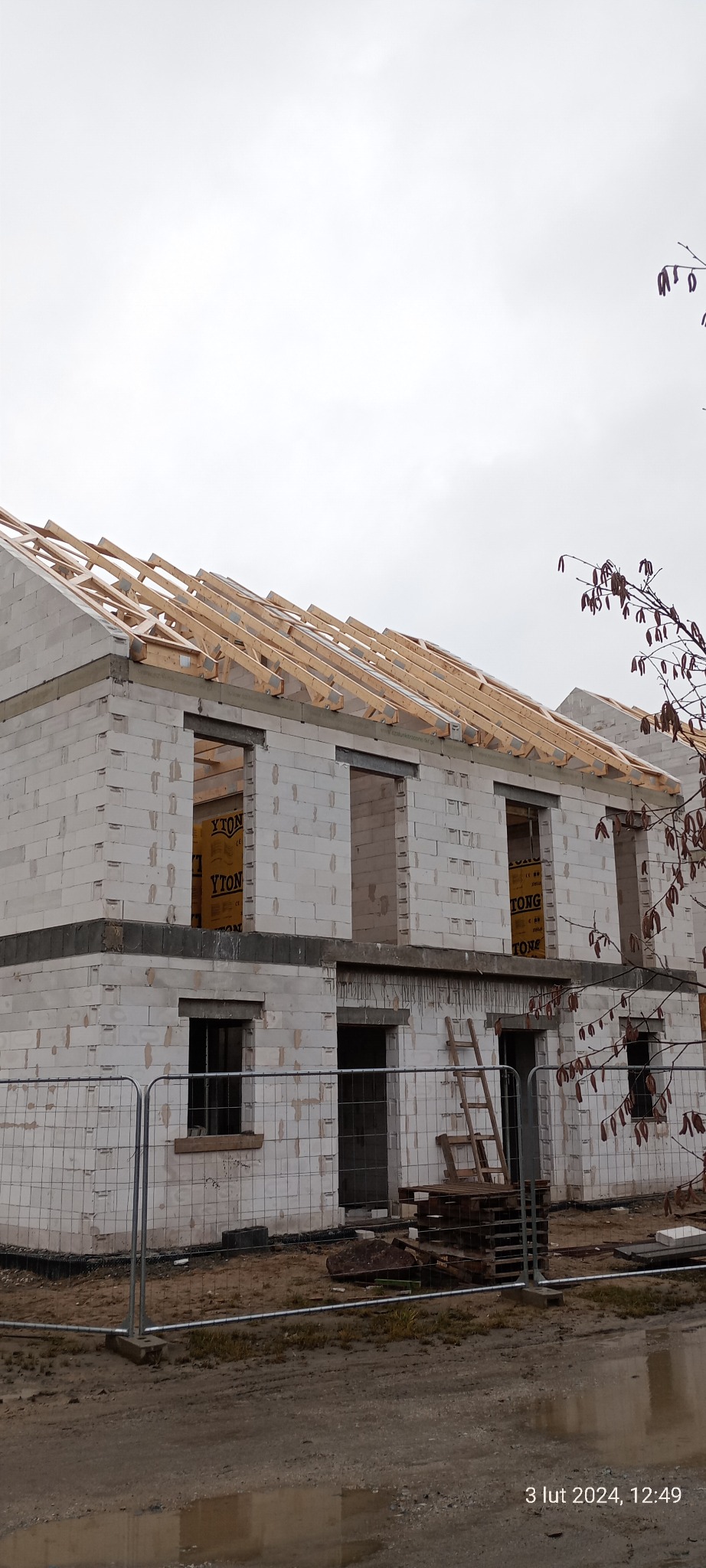 Dwukondygnacyjny budynek w stanie surowym otwartym, wzniesiony z białych bloczków, z widoczną konstrukcją dachu z drewnianych krokwi, zabezpieczony siatką ogrodzeniową.