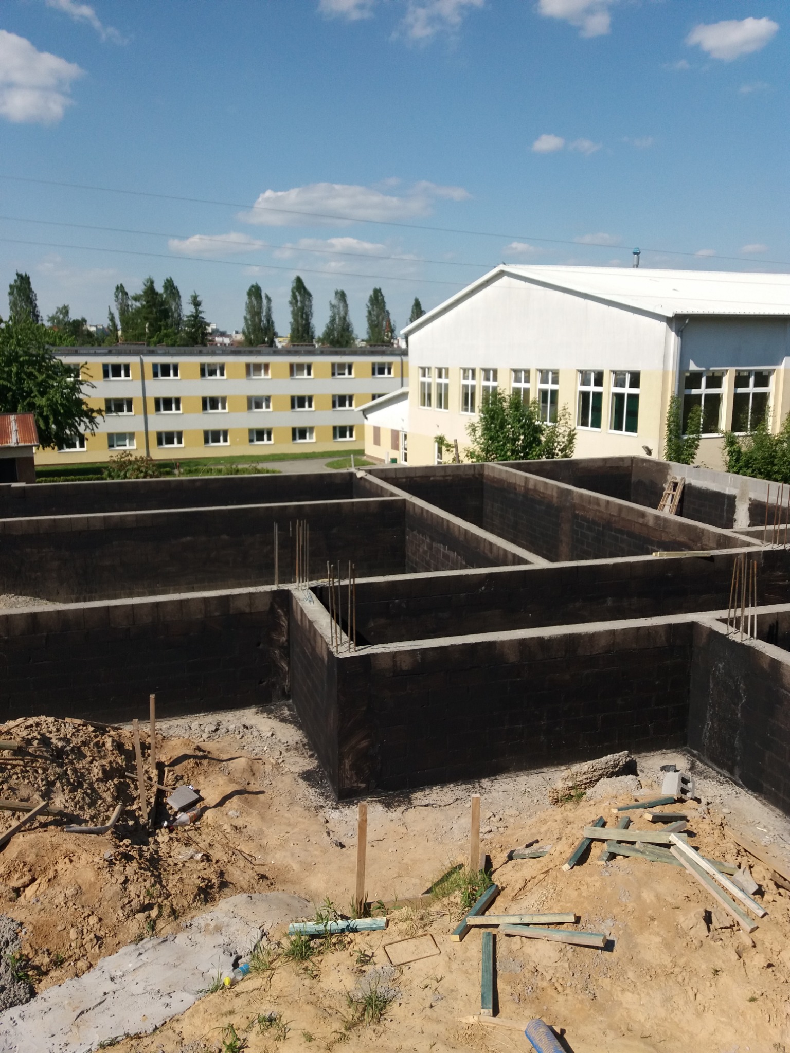 Wykopane i wylane fundamenty budynku z widoczną konstrukcją ścian fundamentowych, zbrojeniami i izolacją przeciwwilgociową. W tle budynki i błękitne niebo.