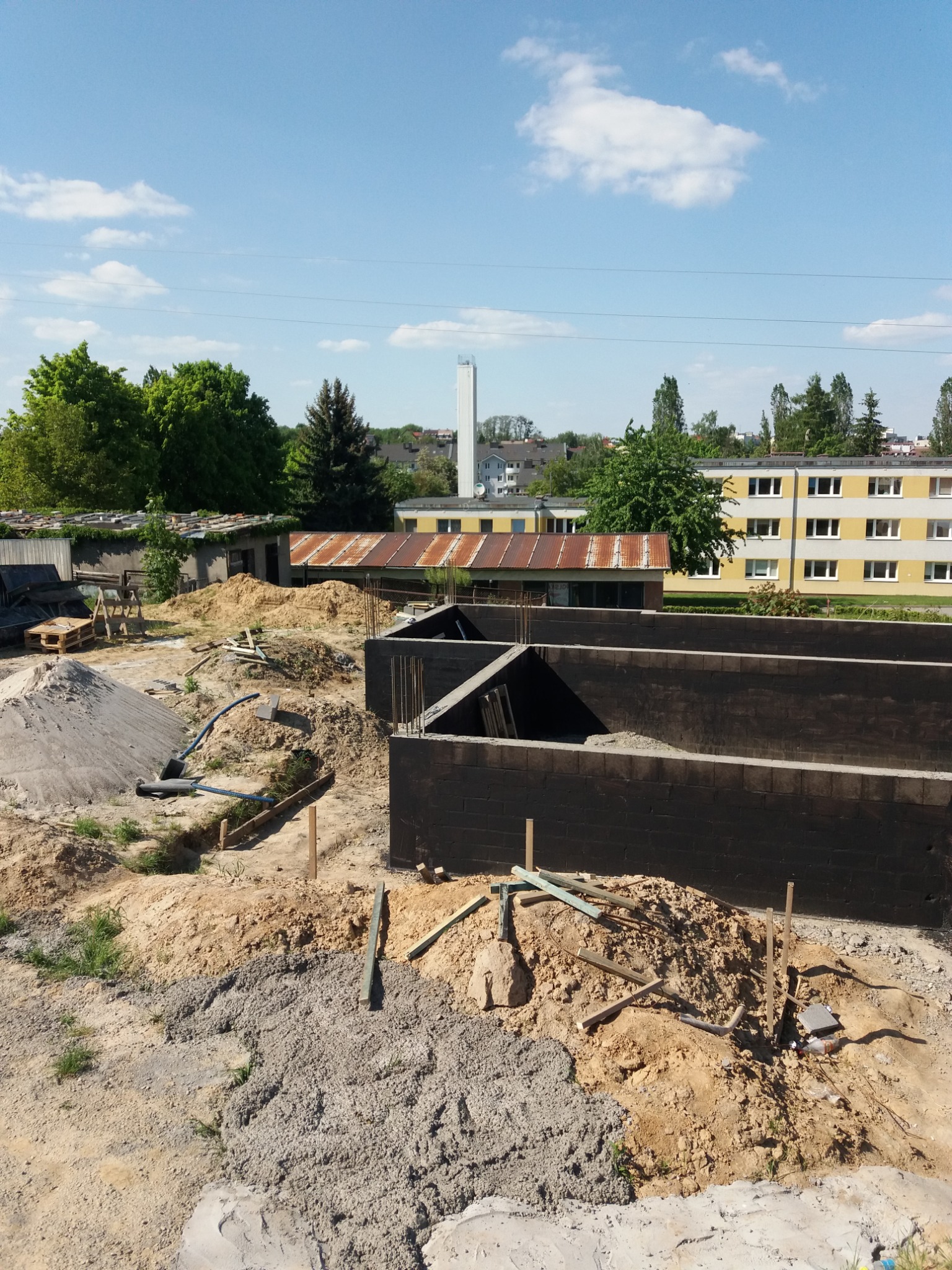 Budowa fundamentów: czarne bloczki fundamentowe zbrojone stalą, ułożone w wykopie, widoczne narzędzia i materiały budowlane, w tle zabudowania miejskie w słoneczny dzień.