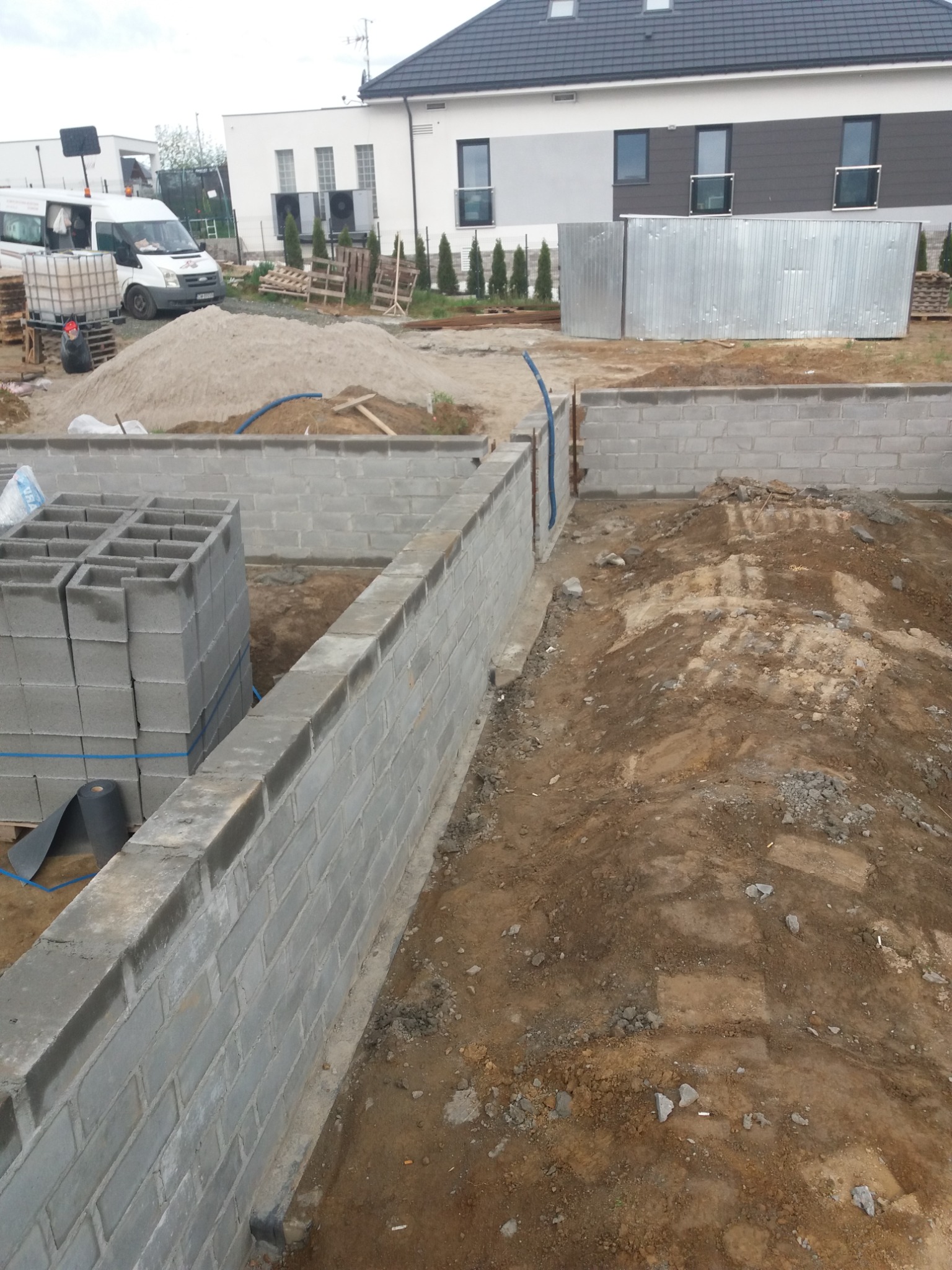 Wznoszenie fundamentów z bloczków betonowych, widoczny stos bloczków na palecie, częściowo wylany fundament, piasek i zaplecze budowy w tle.