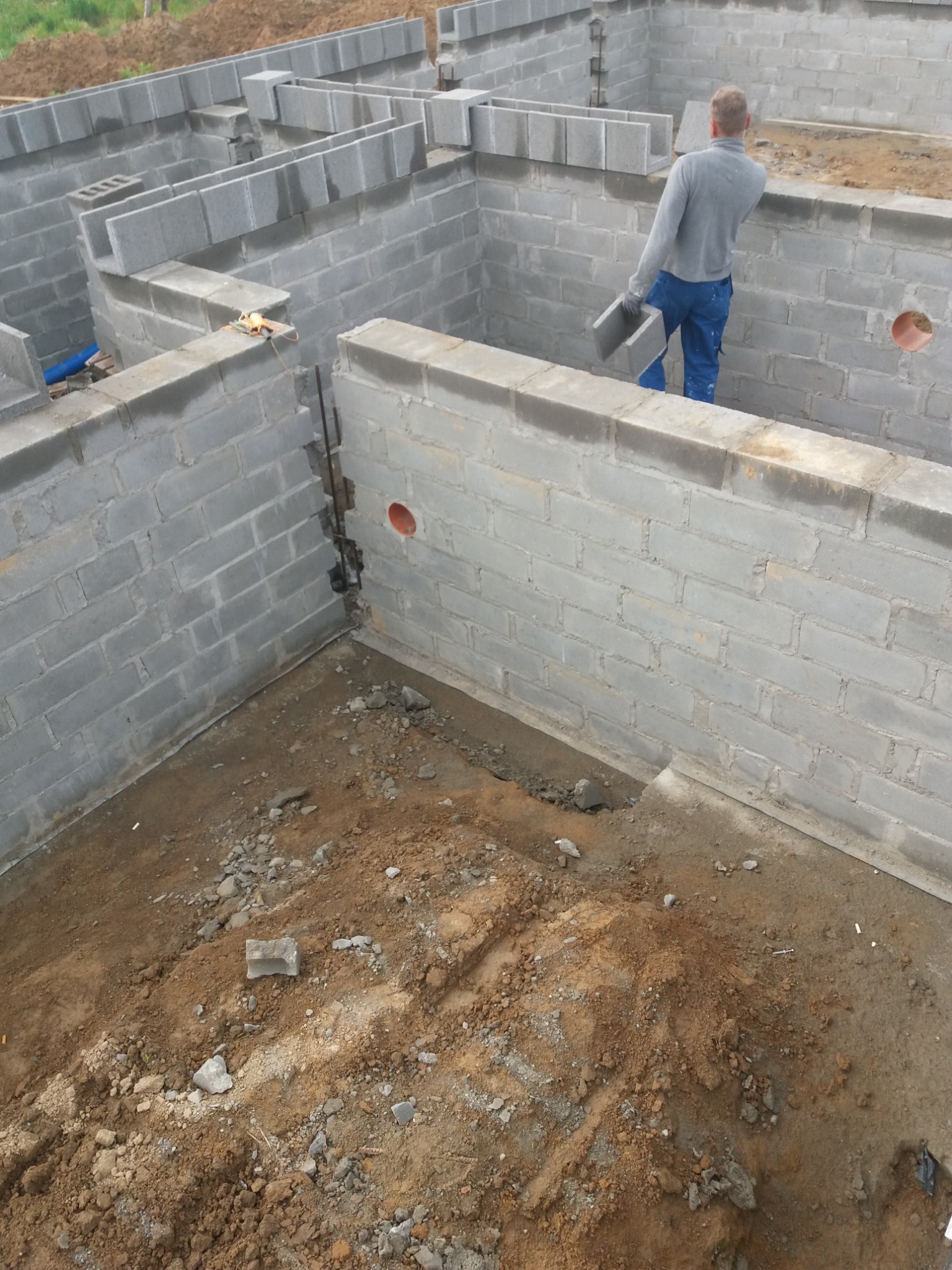 Wznoszenie ścian fundamentowych z szarych bloczków betonowych, widoczny pracownik układający element, przygotowane zbrojenie pionowe, otwór instalacyjny w ścianie, ziemia wewnątrz fundamentów.