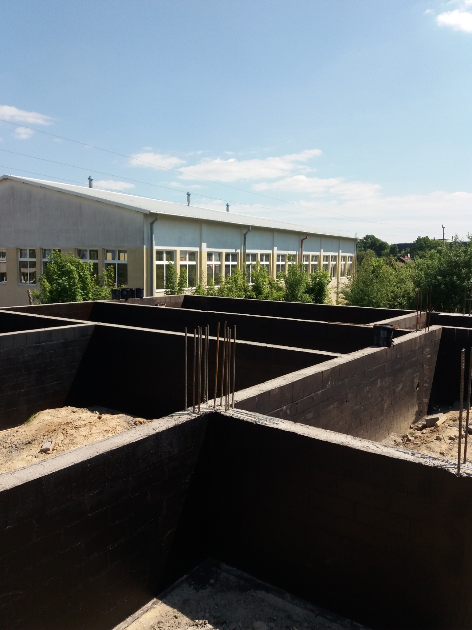 Zalane, czarne fundamenty z wystającymi prętami zbrojeniowymi na tle budynku przemysłowego i błękitnego nieba.