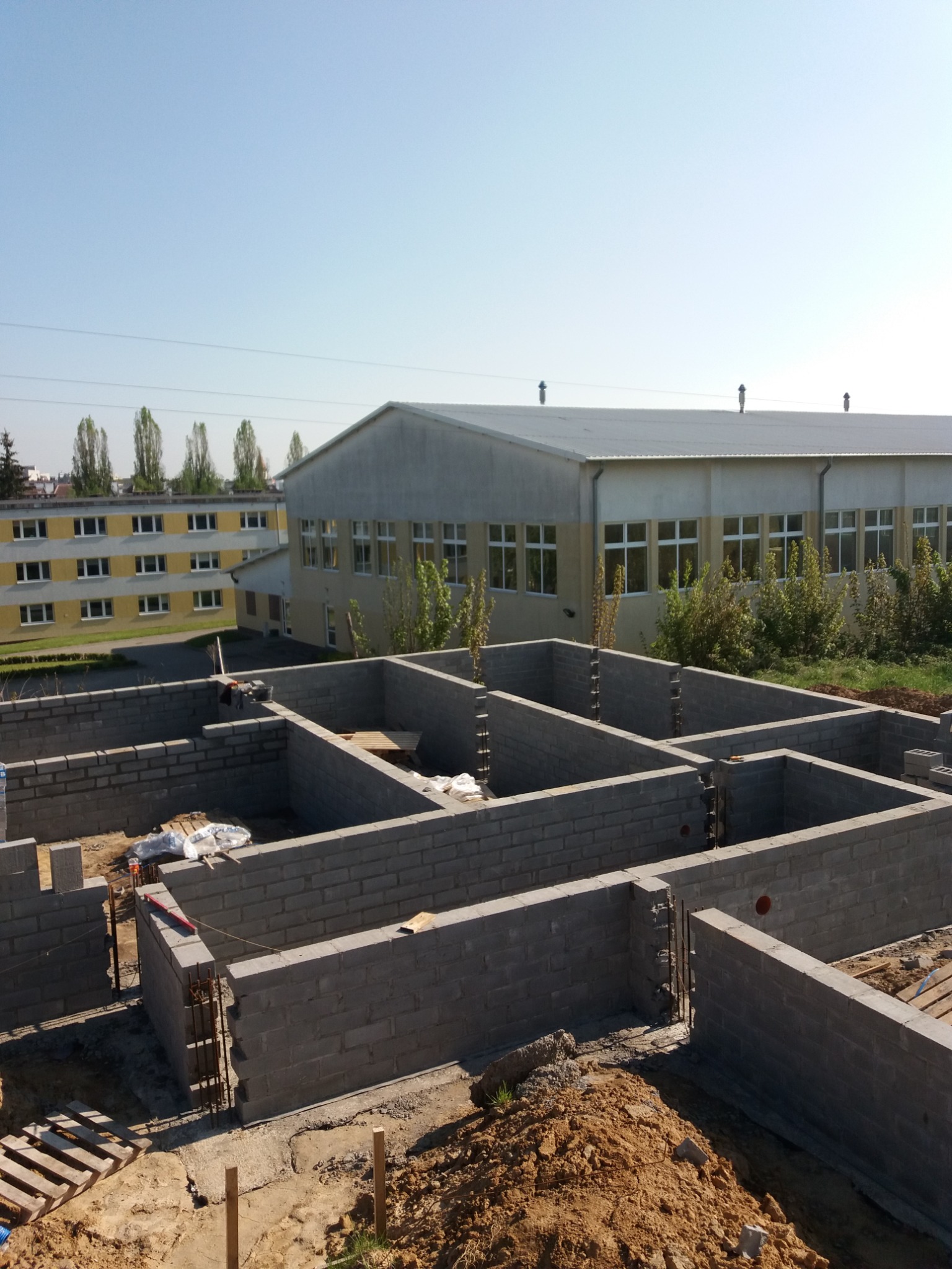 Fundamenty z bloczków betonowych z widocznymi prętami zbrojeniowymi, w tle budynek przemysłowy i drzewa.