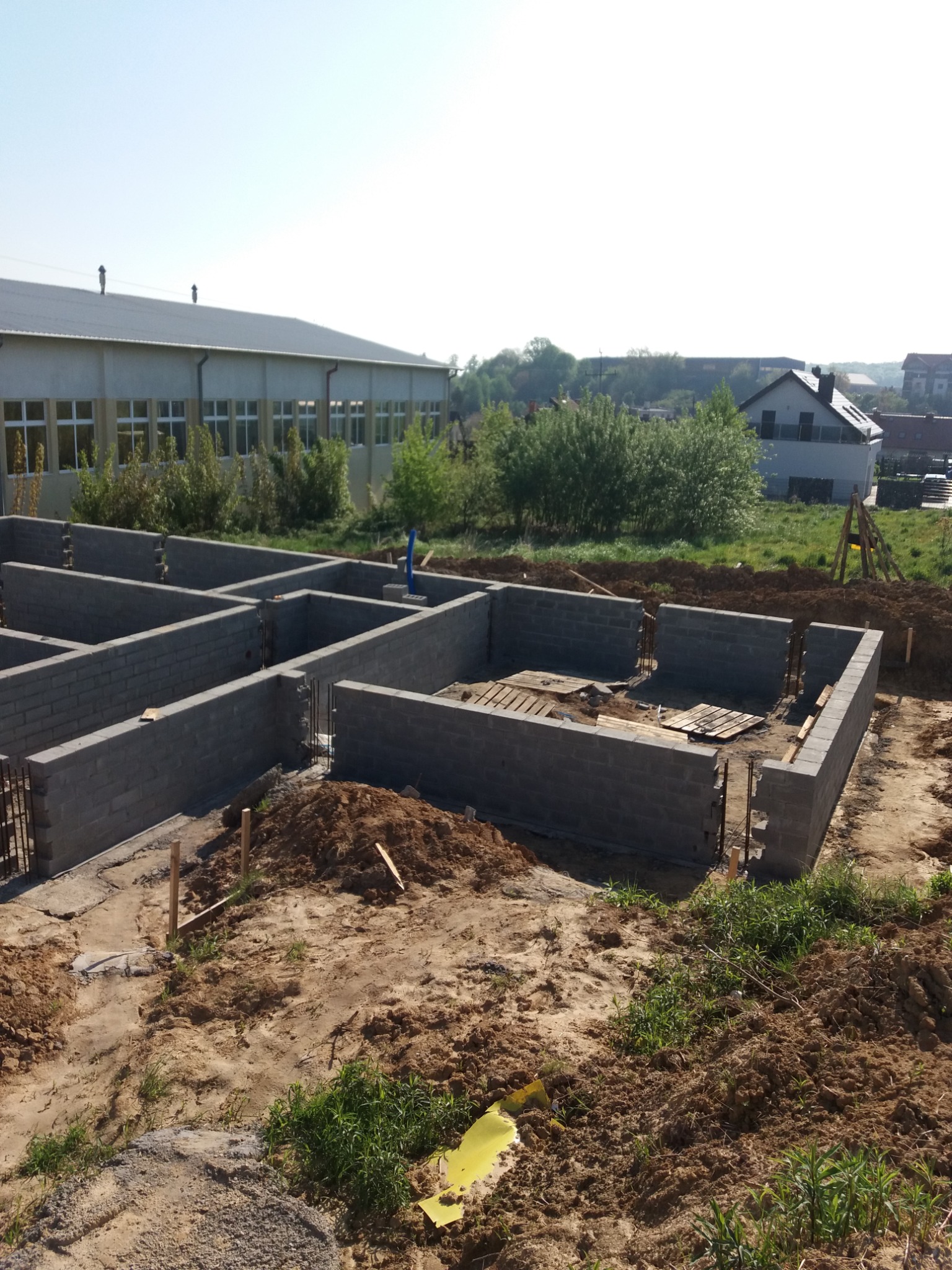Wznoszenie fundamentów z bloczków betonowych, widoczne zbrojenie pionowe i szalunki, teren budowy z ziemią i zielenią w tle, budynek gospodarczy i dom jednorodzinny w oddali.