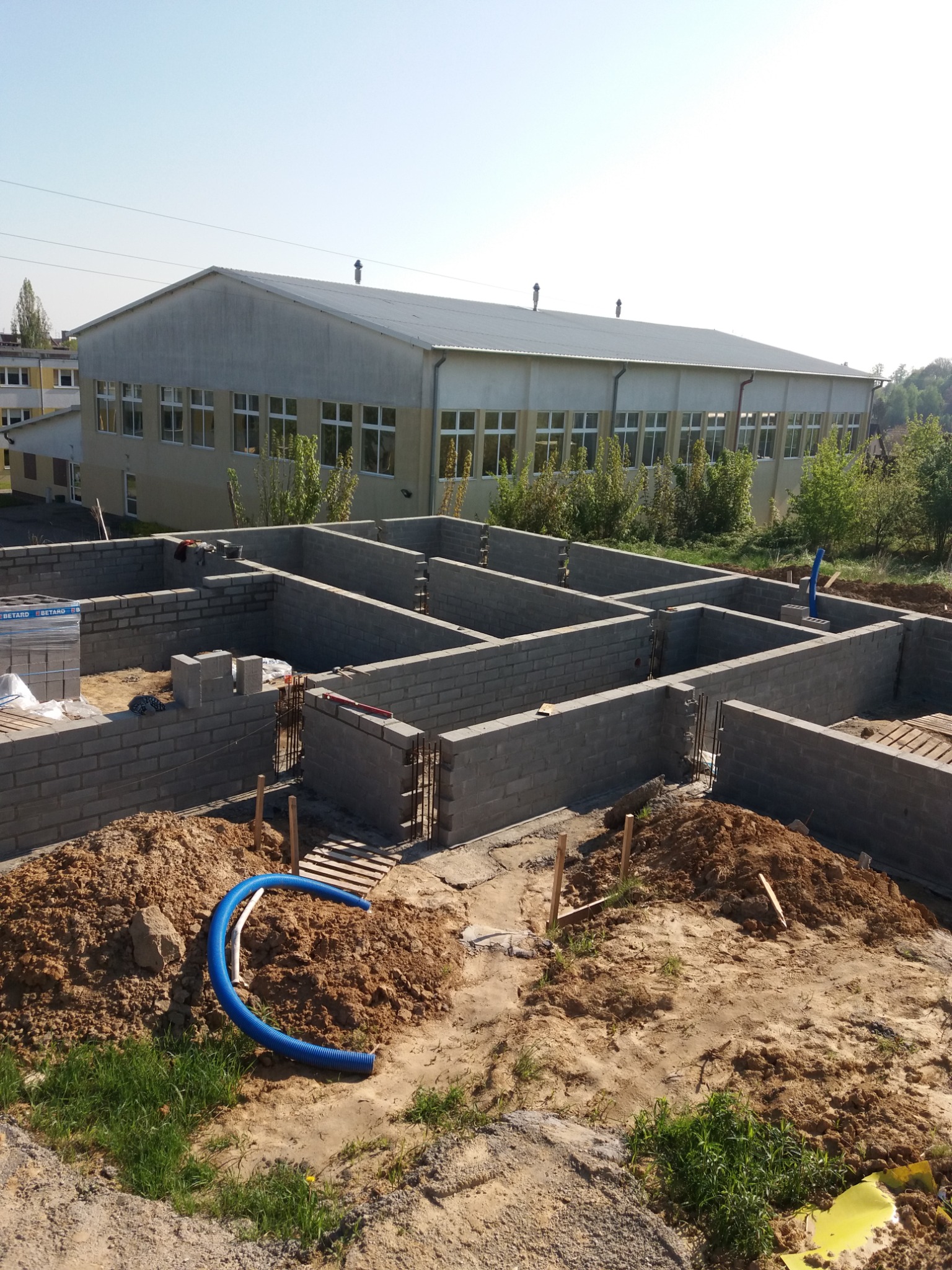 Wykop pod fundamenty budynku z częściowo wzniesionymi ścianami z bloczków betonowych, widoczny niebieski karbowany przewód instalacyjny, w tle budynek gospodarczy.