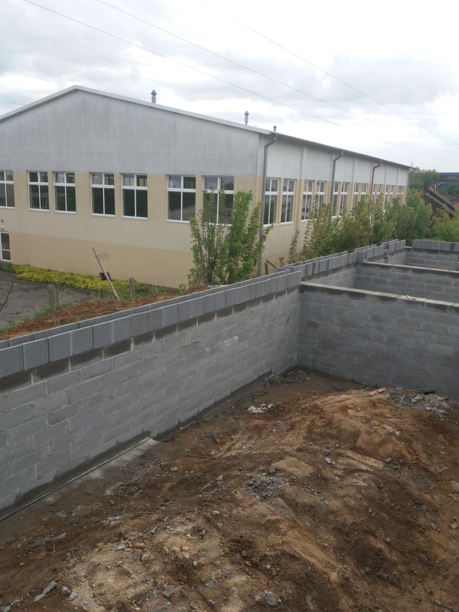 Wykop pod budynek z częściowo wymurowanymi ścianami fundamentowymi z bloczków betonowych, widoczne zbrojenie i zaprawa, w tle budynek gospodarczy.