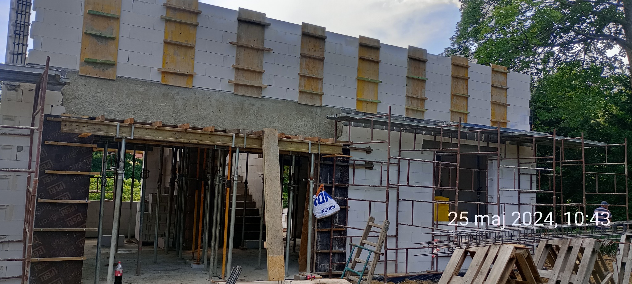 Wznoszenie ścian domu z betonu komórkowego, widoczne szalunki, rusztowania i podpory tymczasowe, teren budowy w otoczeniu drzew.
