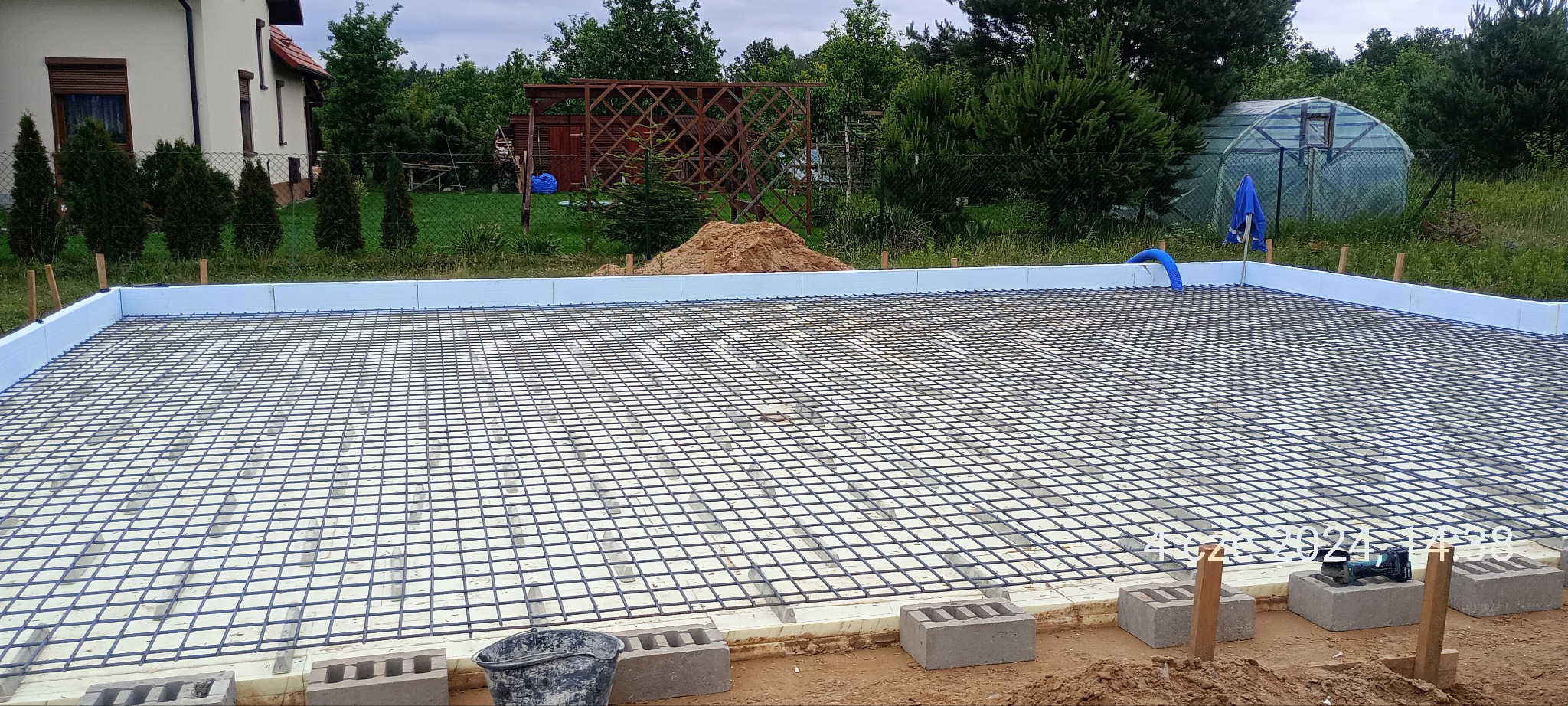 Przygotowany fundament z izolacją styropianową i siatką zbrojeniową, widoczne bloczki betonowe, w tle ogród z tunelem foliowym.