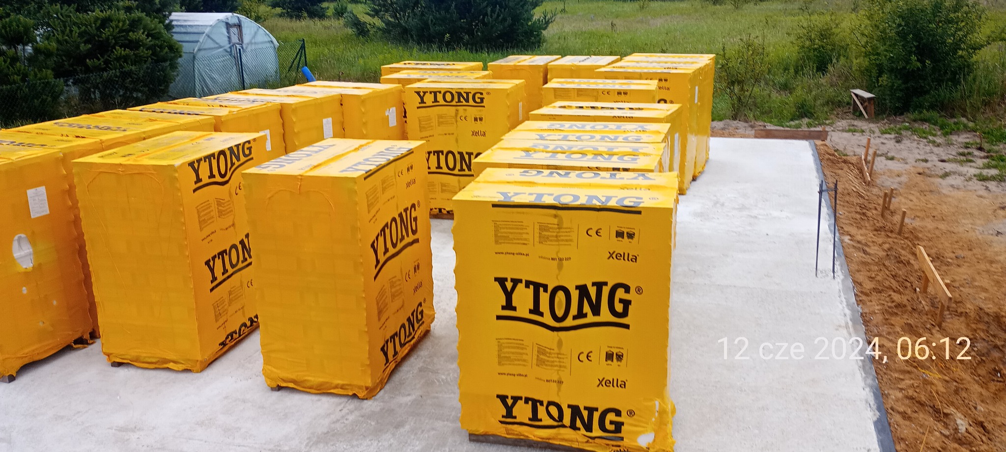 Palety z bloczkami Ytong na płycie fundamentowej, widoczne oznaczenia producenta, w tle zieleń i szklarnia.