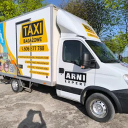 Biały samochód dostawczy z reklamą usług transportowych i przeprowadzkowych, z numerem telefonu i logiem firmy, zaparkowany na placu w słoneczny dzień.