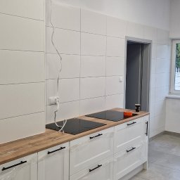 Mar-Greg - Wnętrze kuchni w trakcie wykańczania: białe płytki na ścianach, drewnopodobny blat, białe szafki z czarnymi uchwytami jeszcze w folii ochronnej, płyta indukcyjna, drabina oparta o ścianę przy oknie.