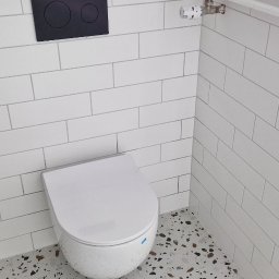 Mar-Greg - Nowoczesna toaleta podwieszana z czarnym panelem sterującym na białych, prostokątnych płytkach ściennych, uzupełniona grzejnikiem drabinkowym i podłogą z lastryko.