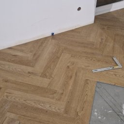 Mar-Greg - Układanie paneli podłogowych w jodełkę; widoczne narzędzia: gumowy młotek, kątownik, kliny dystansowe przy ścianie i fragment surowej posadzki przed położeniem paneli.