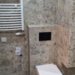 Mar-Greg - Nowoczesna toaleta podwieszana z czarnym panelem sterującym na tle ściany wyłożonej płytkami imitującymi marmur, obok grzejnik drabinkowy.