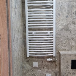 Mar-Greg - Biały grzejnik drabinkowy zamontowany na ścianie z płytek imitujących kamień w łazience, widoczny fragment drewnianych drzwi i osprzęt instalacyjny.