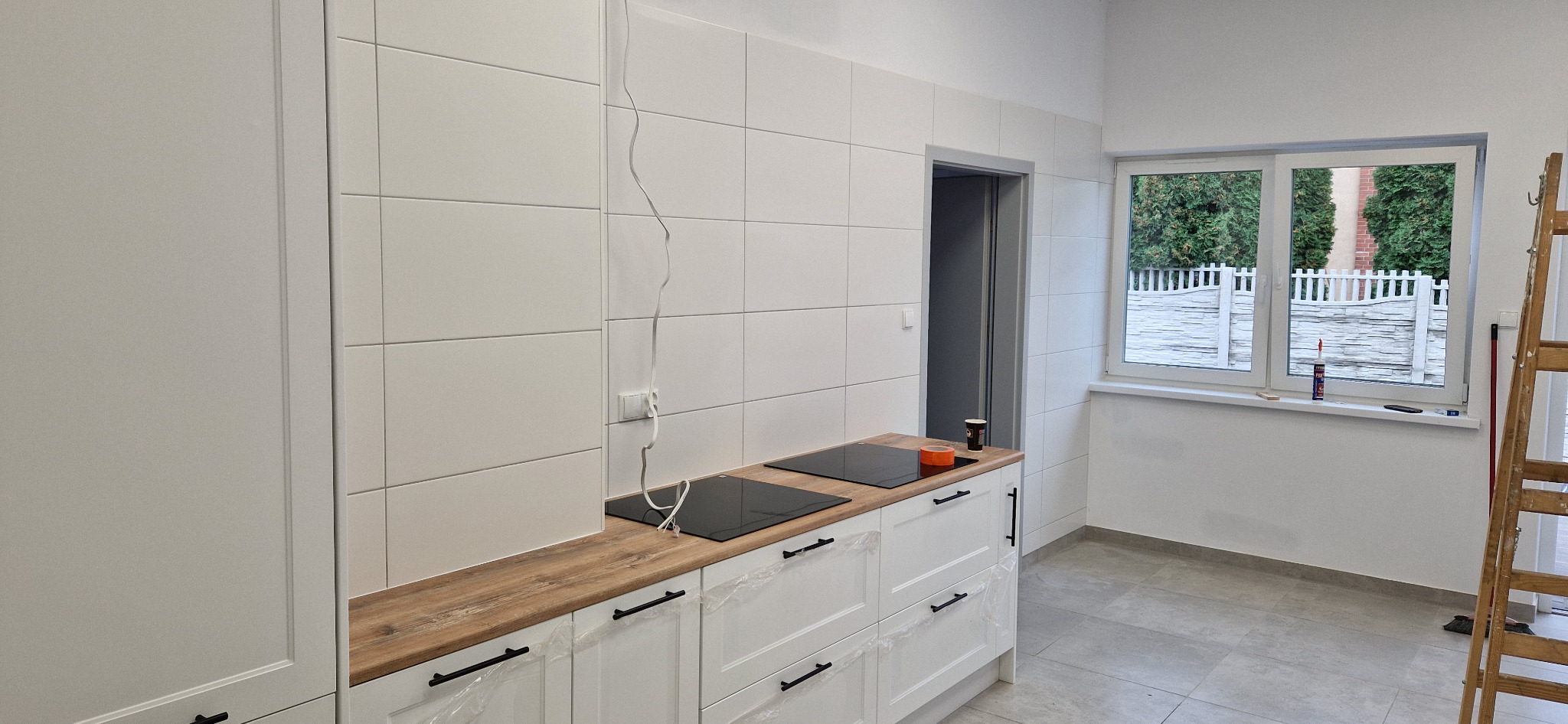 Wnętrze kuchni w trakcie wykańczania: białe płytki na ścianach, drewnopodobny blat, białe szafki z czarnymi uchwytami jeszcze w folii ochronnej, płyta indukcyjna, drabina oparta o ścianę przy oknie.