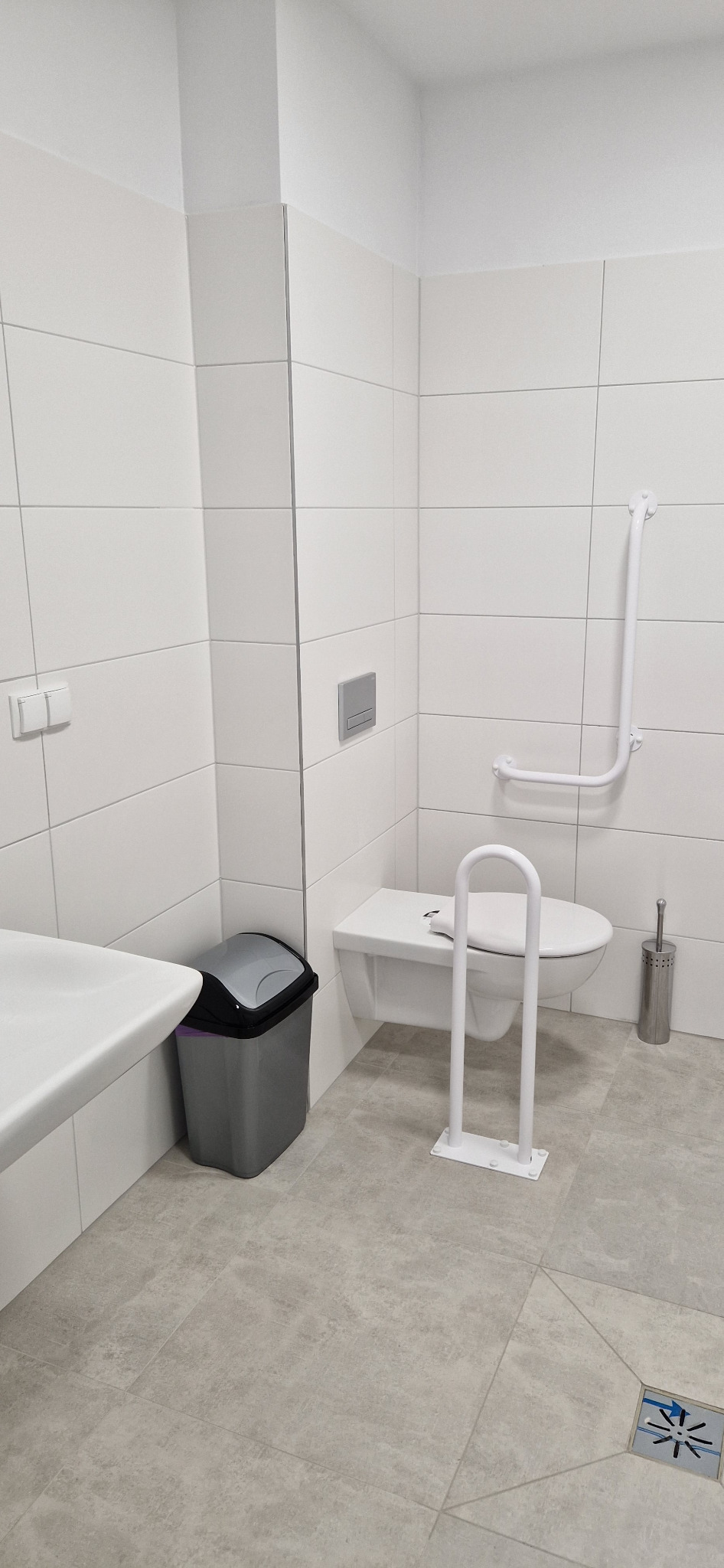 Nowoczesna łazienka z białą armaturą, płytkami w odcieniach szarości i uchwytami dla osób z niepełnosprawnościami, widok na toaletę z dodatkowym wsparciem, umywalkę i kosz na śmieci.