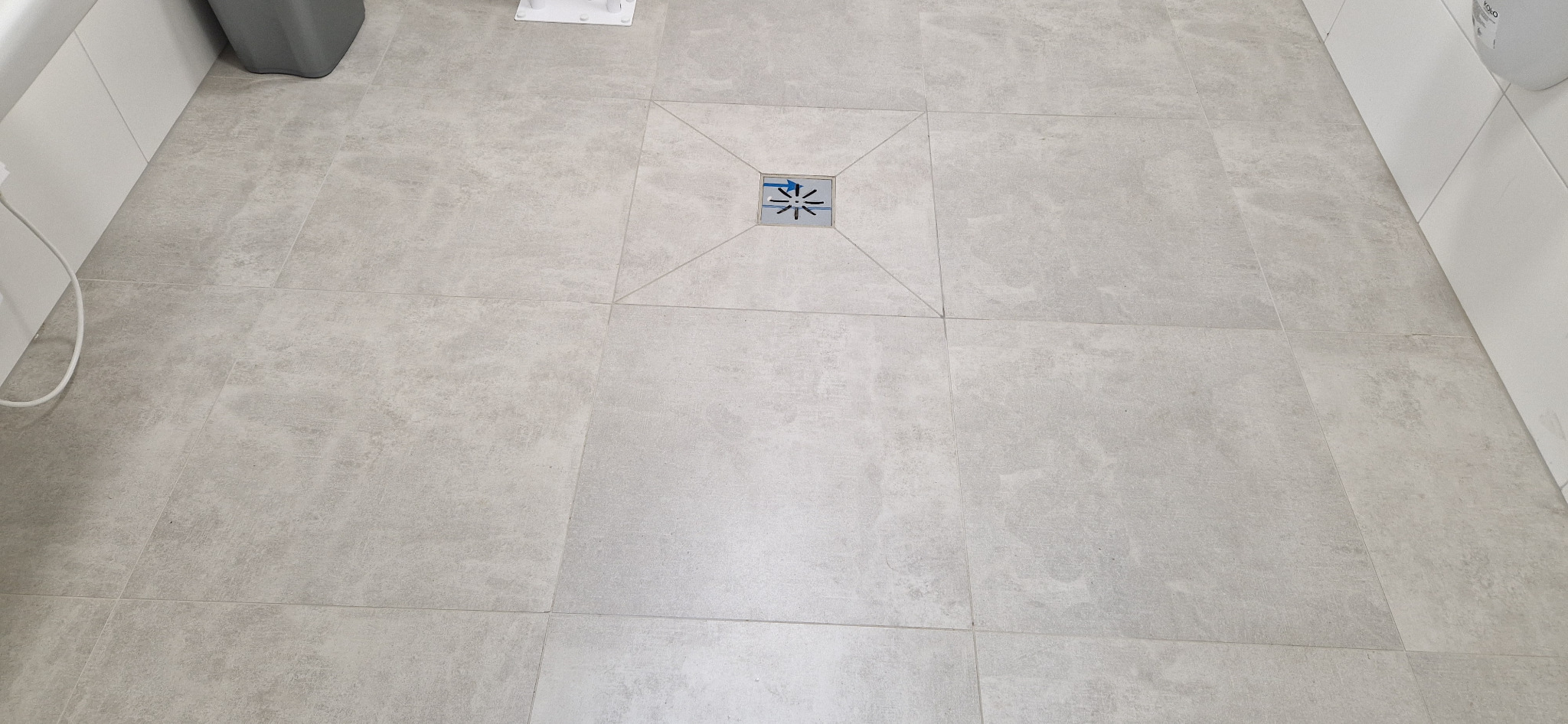 Jasnoszara, matowa podłoga z płytek ceramicznych z centralnie umieszczonym odpływem liniowym w kwadratowej ramce, widok z góry.