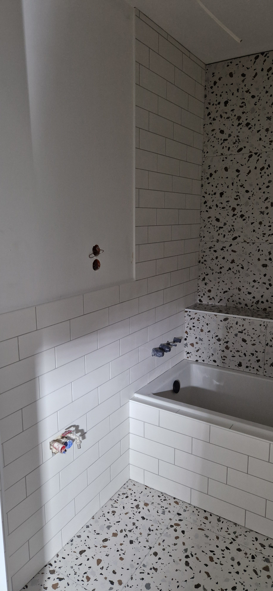 Biała łazienka w trakcie remontu: ściany wyłożone prostokątnymi płytkami, fragment ściany z terazzo, widoczne rury i zawory przed montażem armatury, surowy stan wykończenia.