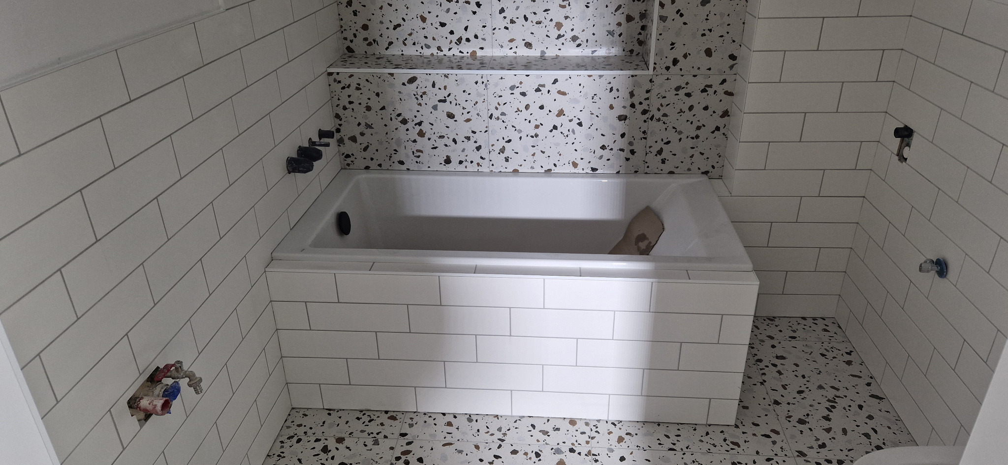 Wanna obudowana białymi, prostokątnymi płytkami, ściany wyłożone białymi płytkami typu metro, fragment ściany i podłogi z płytkami lastryko, widoczne czarne krany i niedokończone podłączenie wodne.