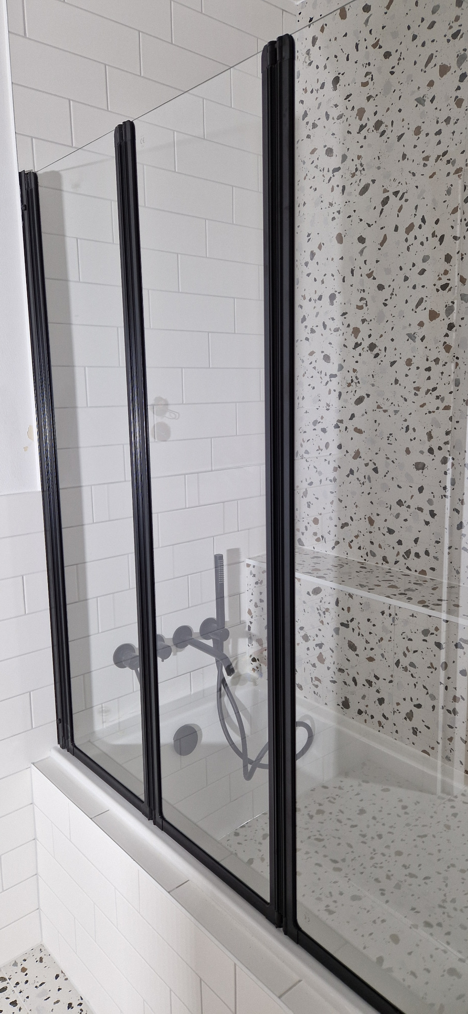 Szklana kabina prysznicowa z czarnymi profilami, białe płytki typu metro na ścianie i mozaika lastryko wewnątrz kabiny, bateria podtynkowa w kolorze czarnym.