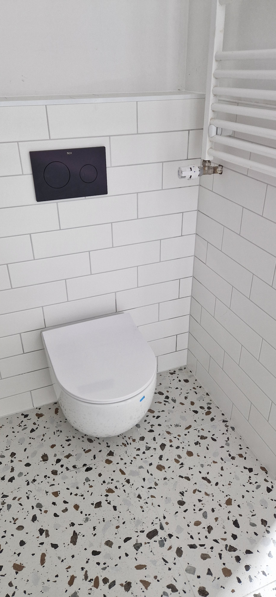 Nowoczesna toaleta podwieszana z czarnym panelem sterującym na białych, prostokątnych płytkach ściennych, uzupełniona grzejnikiem drabinkowym i podłogą z lastryko.