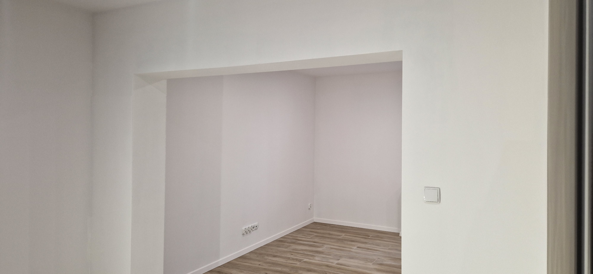 Pomalowane na biało wnętrze z drewnopodobną podłogą, widoczne gniazdka i włącznik światła, przygotowane do dalszego wykończenia lub aranżacji.