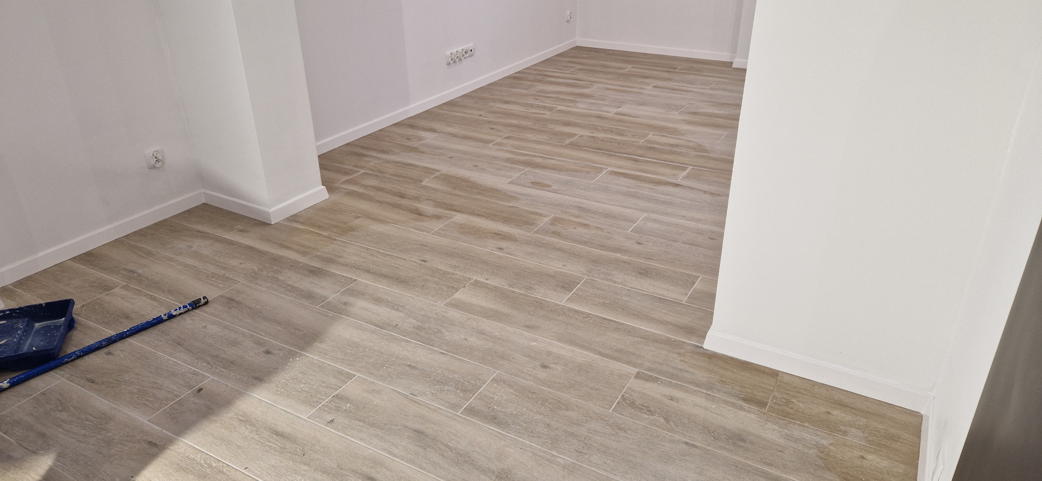 Ułożona podłoga z płytek imitujących drewno w pustym, świeżo wykończonym pomieszczeniu z białymi ścianami i listwami przypodłogowymi, widoczne narzędzia malarskie w rogu.