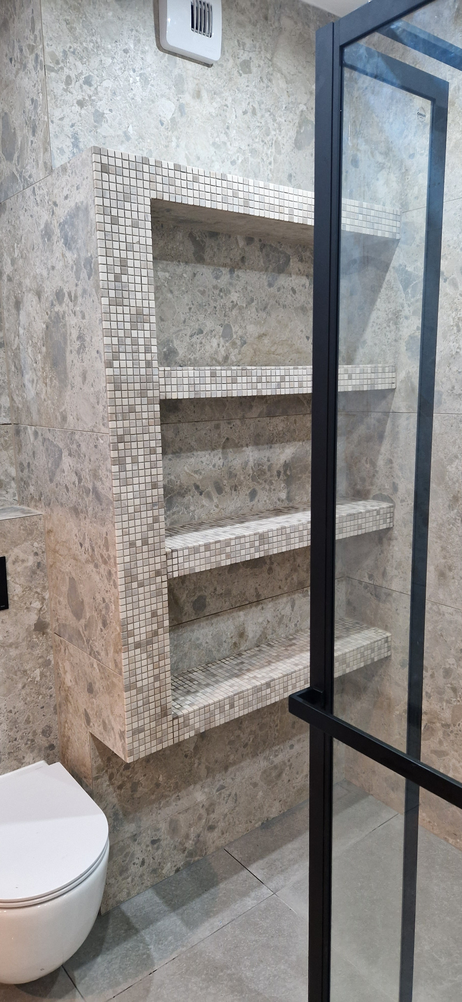 Łazienka z marmurowymi płytkami, wbudowane półki wykończone mozaiką, częściowo widoczna kabina prysznicowa z czarnymi profilami i biała toaleta.
