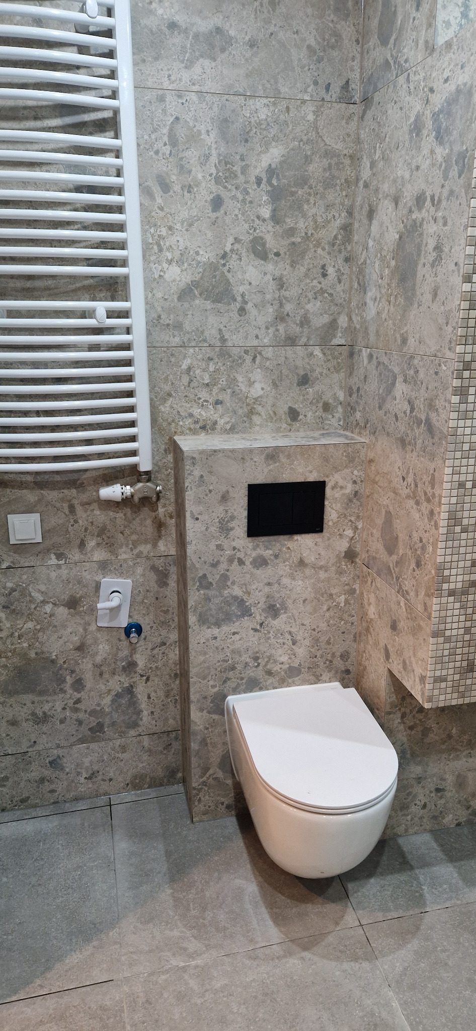 Nowoczesna toaleta podwieszana z czarnym panelem sterującym na tle ściany wyłożonej płytkami imitującymi marmur, obok grzejnik drabinkowy.