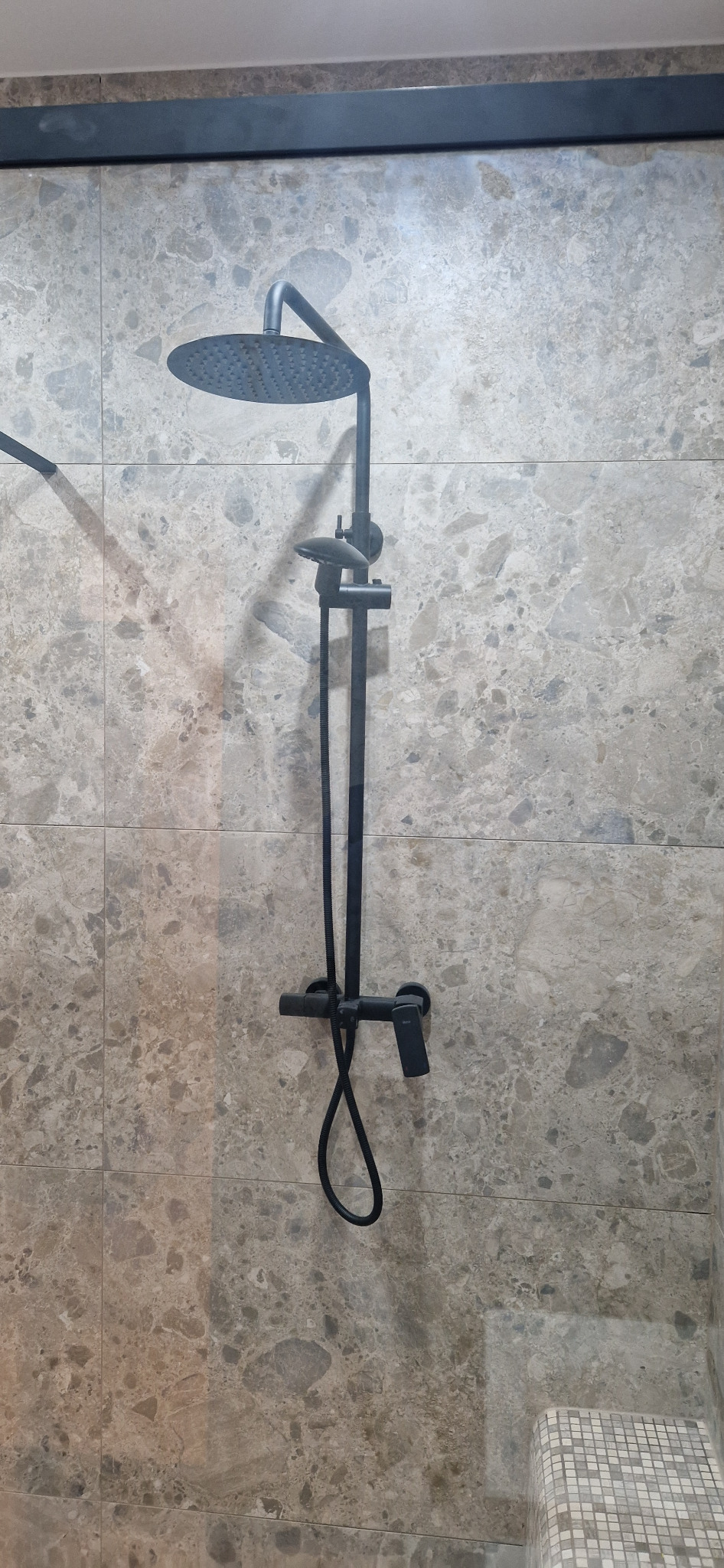 Zamontowany czarny zestaw prysznicowy z deszczownicą na ścianie wyłożonej płytkami imitującymi marmur w odcieniach szarości i beżu, widoczna mozaika podłogowa.