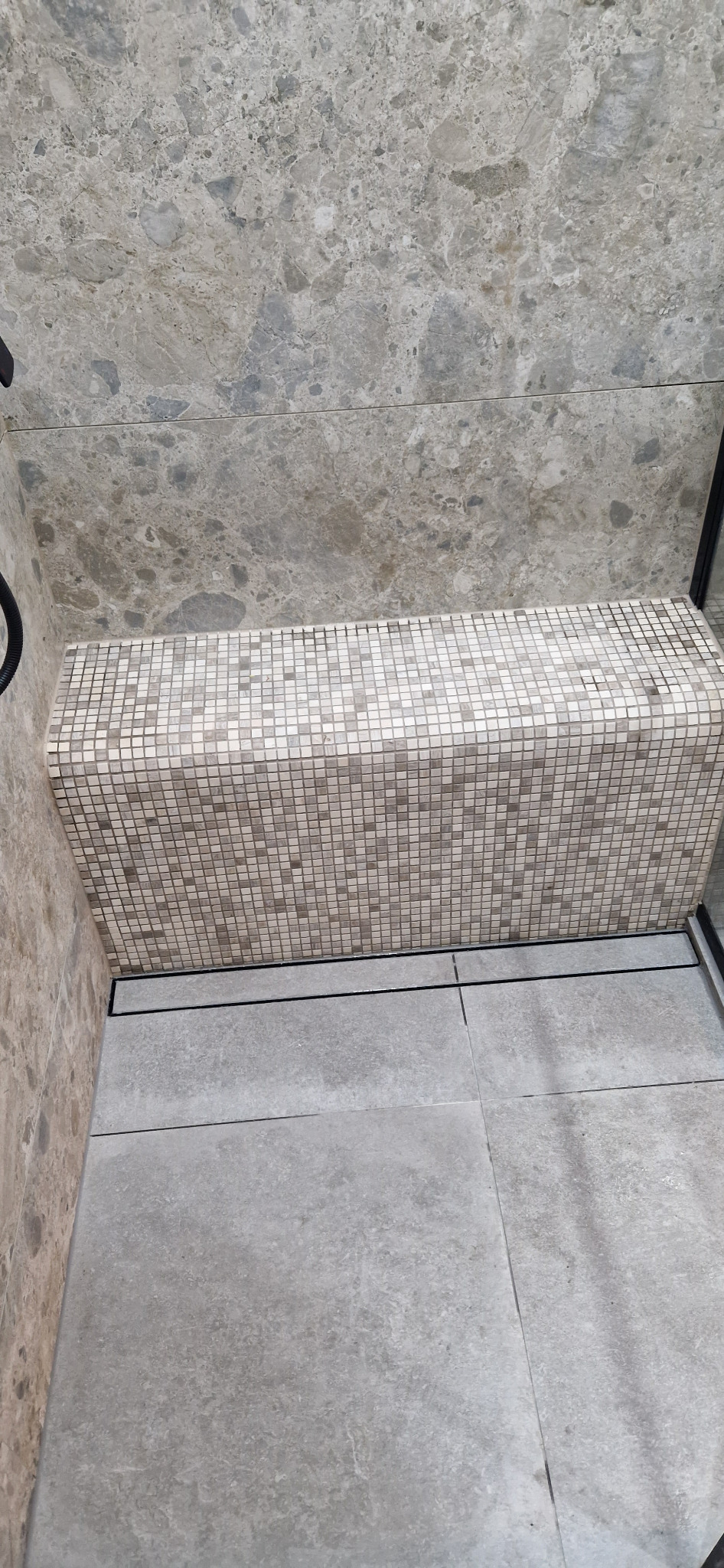 Wnętrze łazienki z siedziskiem wyłożonym mozaiką w strefie prysznicowej, szare płytki podłogowe i ścienne z wzorem kamienia.