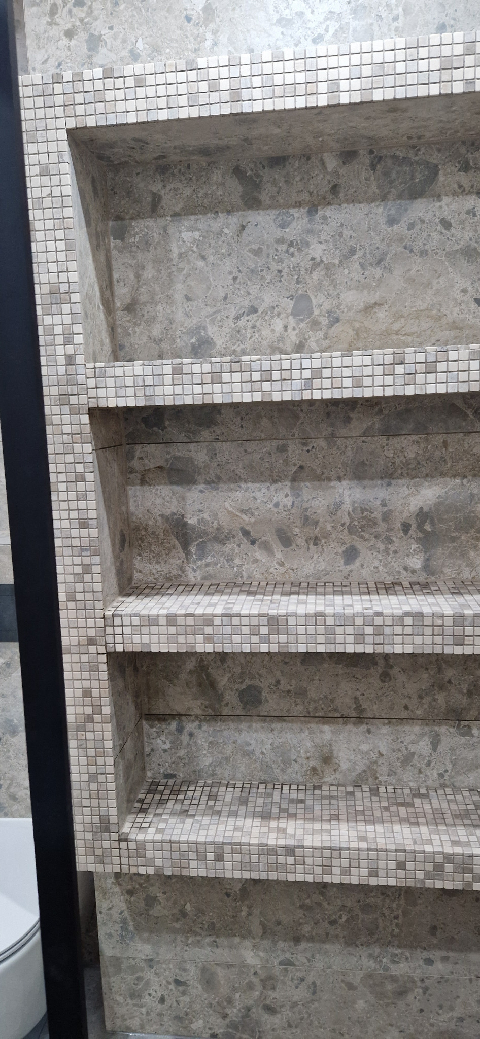 Wnęka w łazience wykończona szarym marmurem i mozaiką z drobnych płytek, z widocznym fragmentem toalety po lewej stronie.